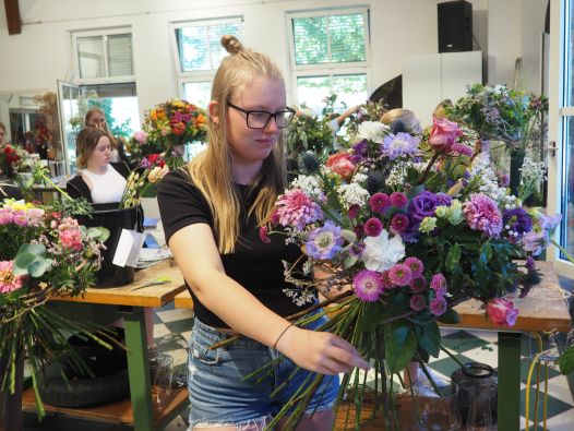 Eine Schülerin begutachtet ihren dekorativen Strauß in den Farben weiß, pink bis Violett, den sie im Praxisunterricht Floristik bindet